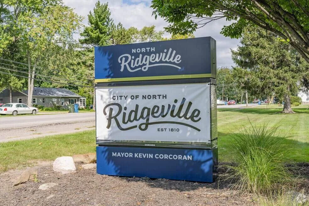 Welcome sign for the City of North Ridgeville, Ohio, featuring the name of Mayor Kevin Corcoran, located near a grassy roadside with trees and houses in the background.