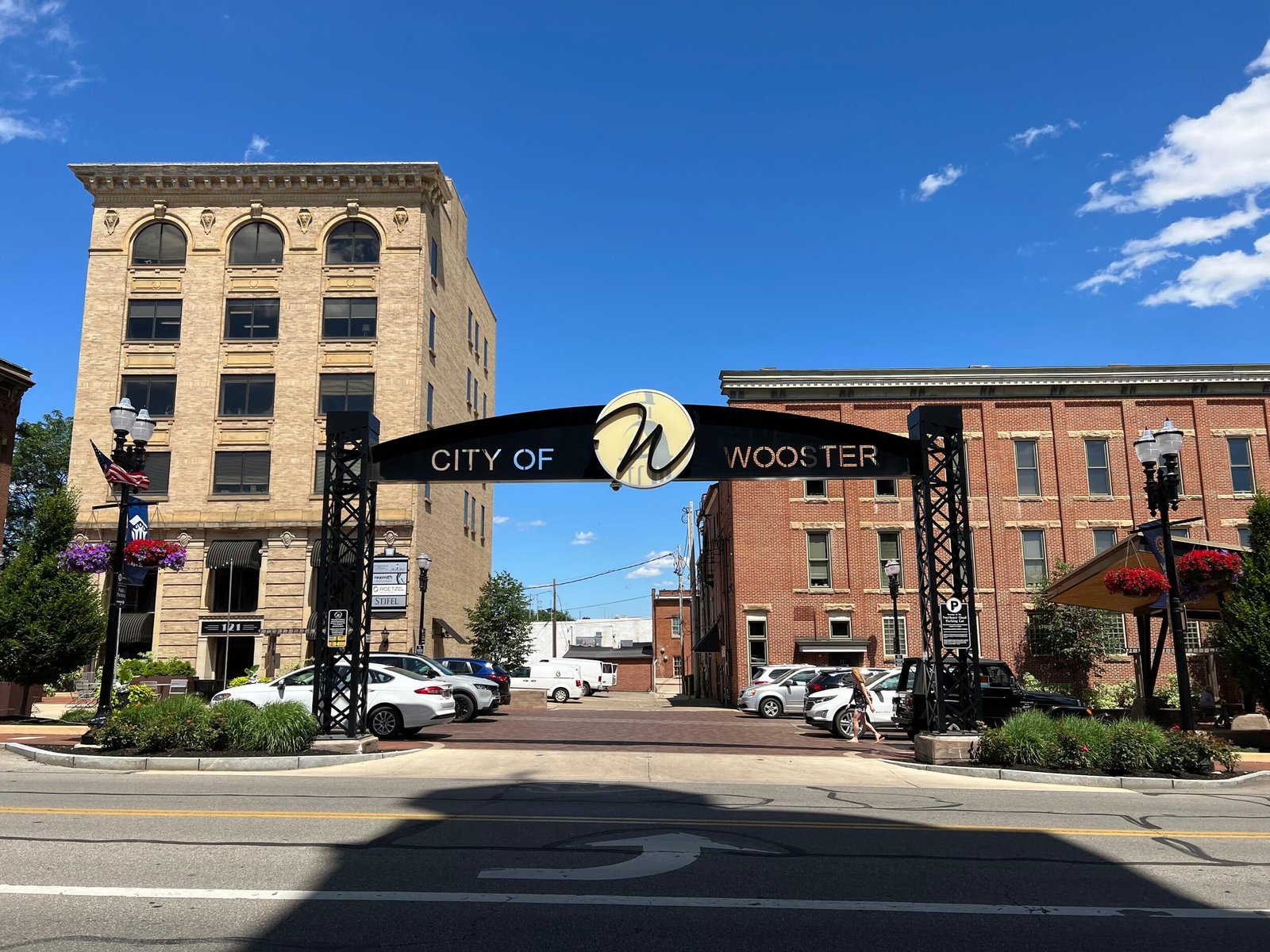 City of Wooster entrance archway with historic brick buildings, parked cars, and hanging flower baskets on a sunny day with clear blue skies.