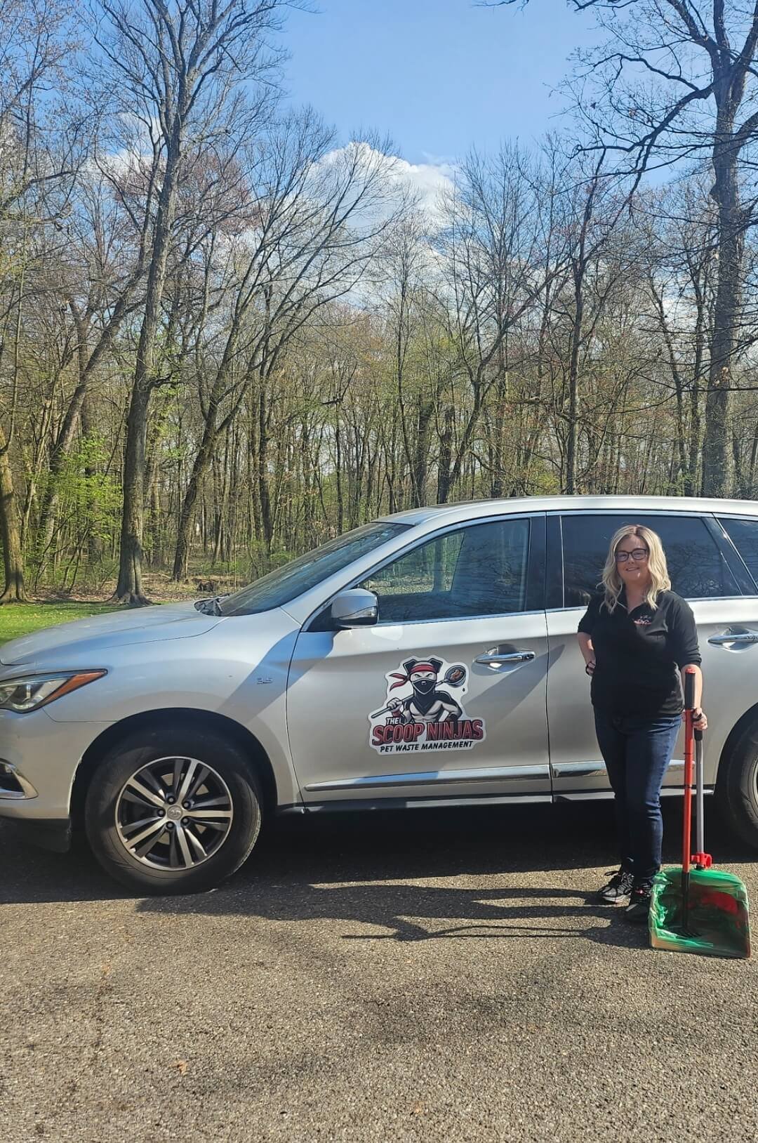Female Scoop Tech from The Scoop Ninjas™ stands beside a branded pet waste management vehicle holding a pooper scooper, ready for service in a wooded residential area.