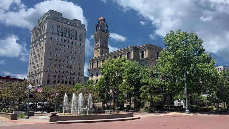 Downtown North Canton, Ohio skyline featuring a historic clock tower, a tall office building, and a central fountain surrounded by trees and American flags under a partly cloudy sky.