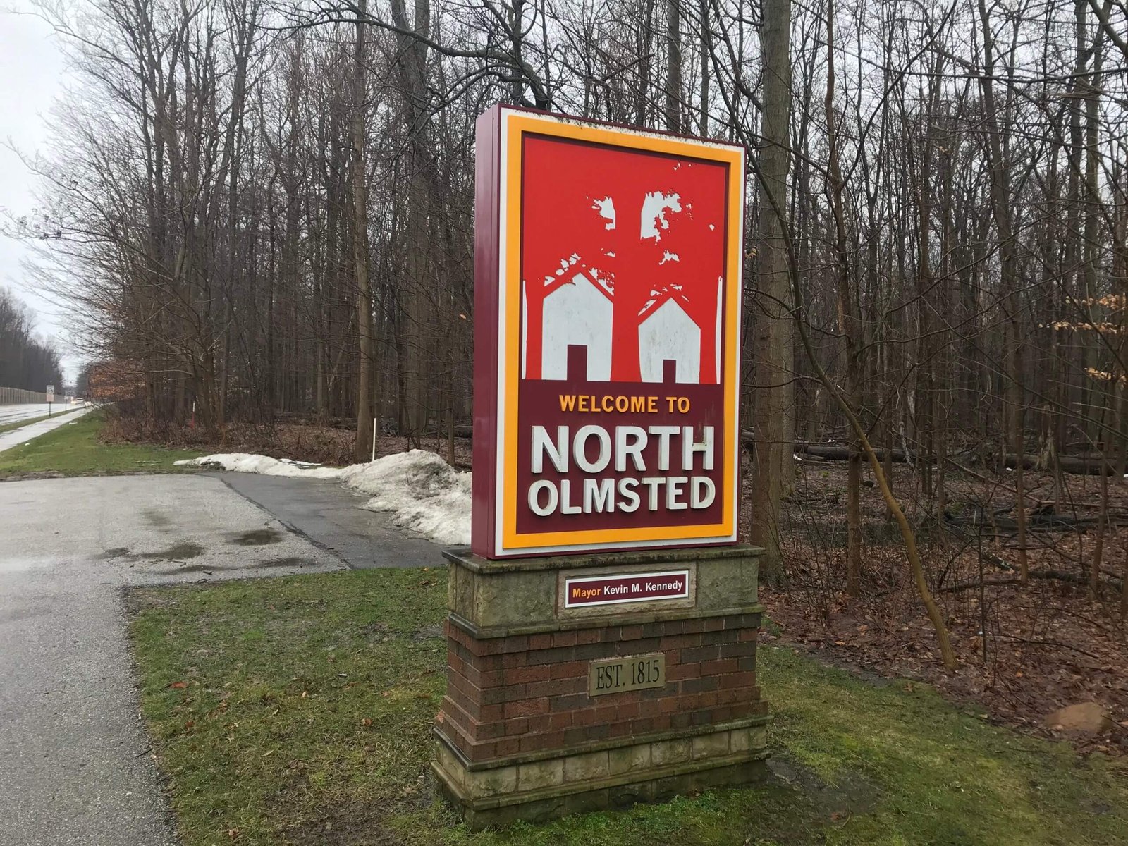 Welcome sign for North Olmsted, Ohio, featuring bold red and white graphics with tree and house silhouettes, set against a wooded background on a cloudy, early spring day.