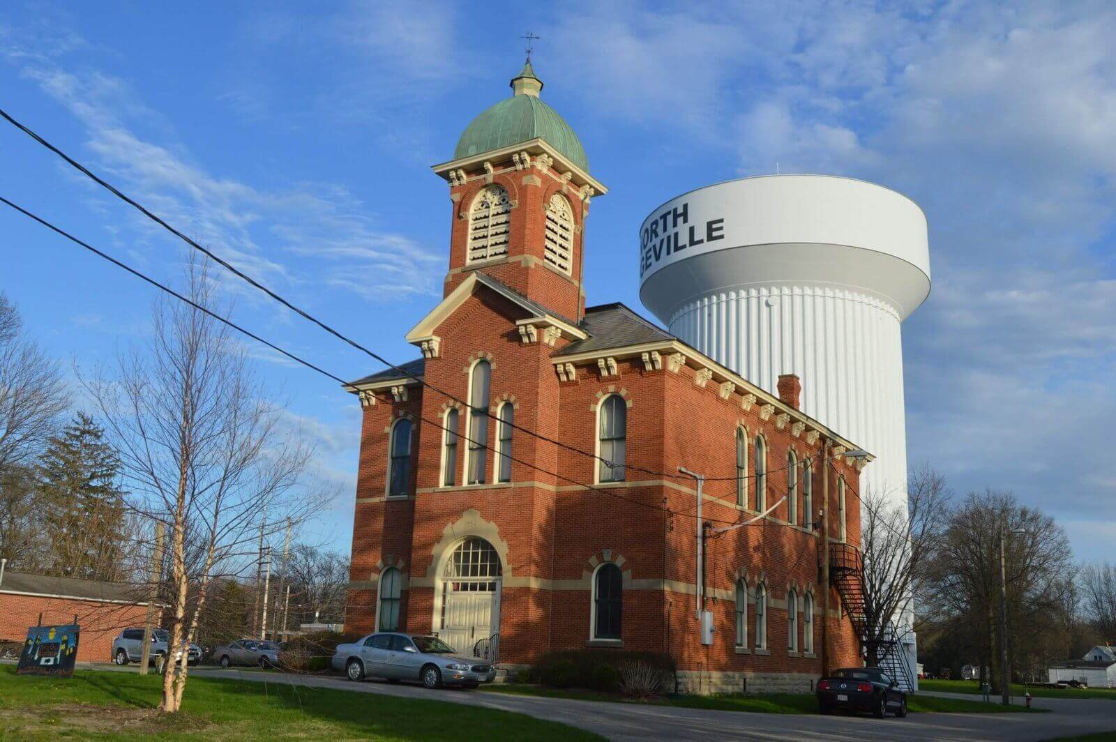 Historic red-brick North Ridgeville City Hall building with a green-domed clock tower, set in front of a large white water tower labeled "North Ridgeville" under a partly cloudy blue sky.