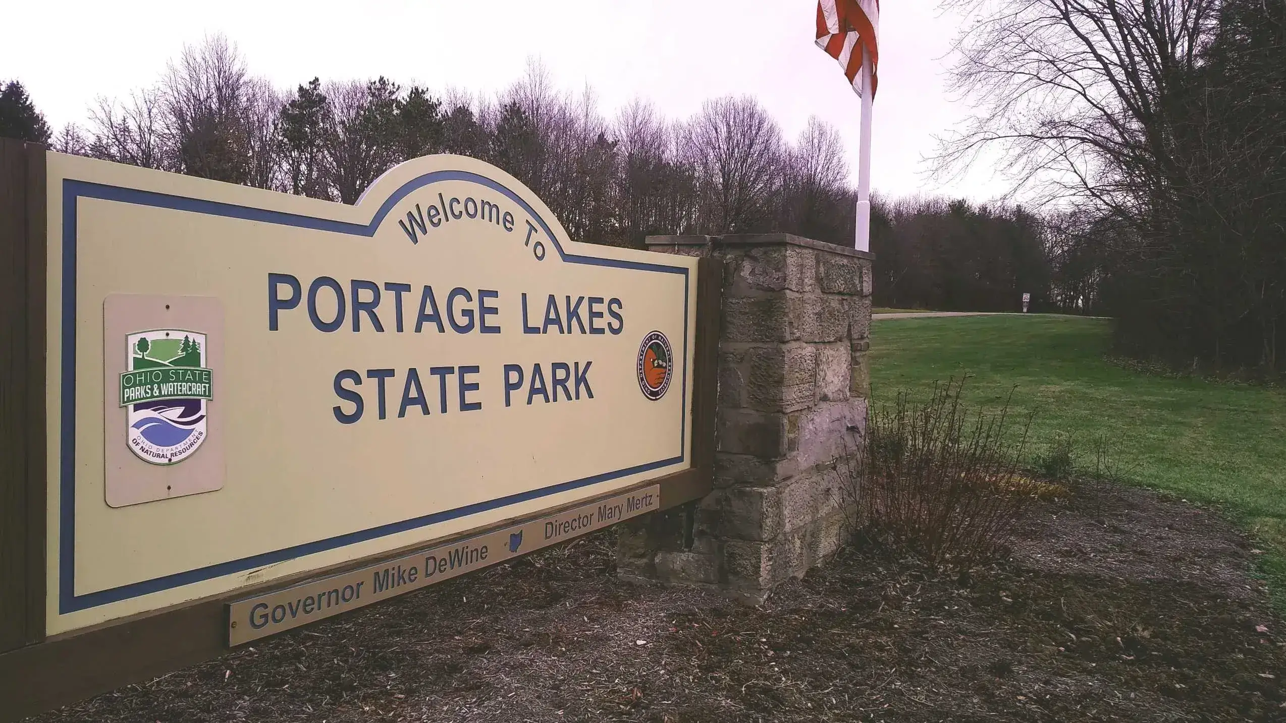 Entrance sign for Portage Lakes State Park in Ohio, featuring the Ohio State Parks & Watercraft logo, set beside a stone pillar and grassy parkland under an overcast sky.