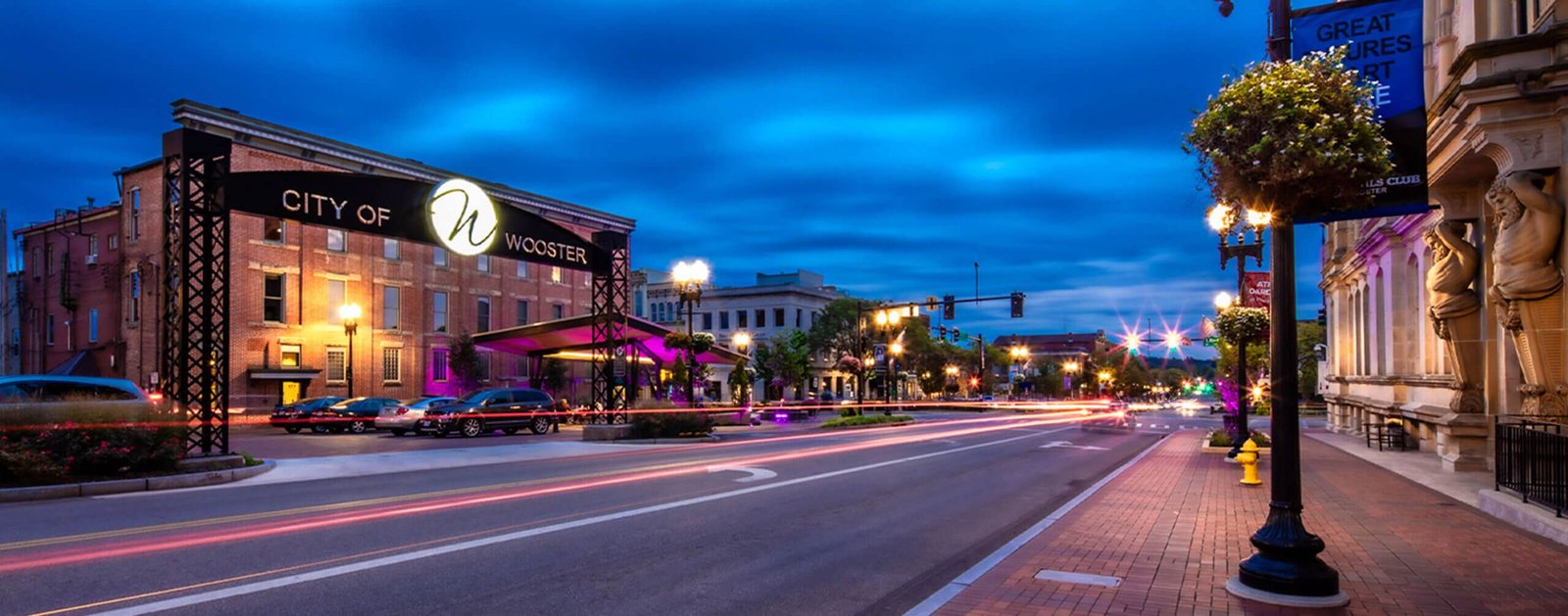 Vibrant nighttime view of downtown Wooster, Ohio, featuring the illuminated “City of Wooster” sign, historic brick buildings, glowing streetlights, and motion-blurred car lights creating a lively atmosphere.